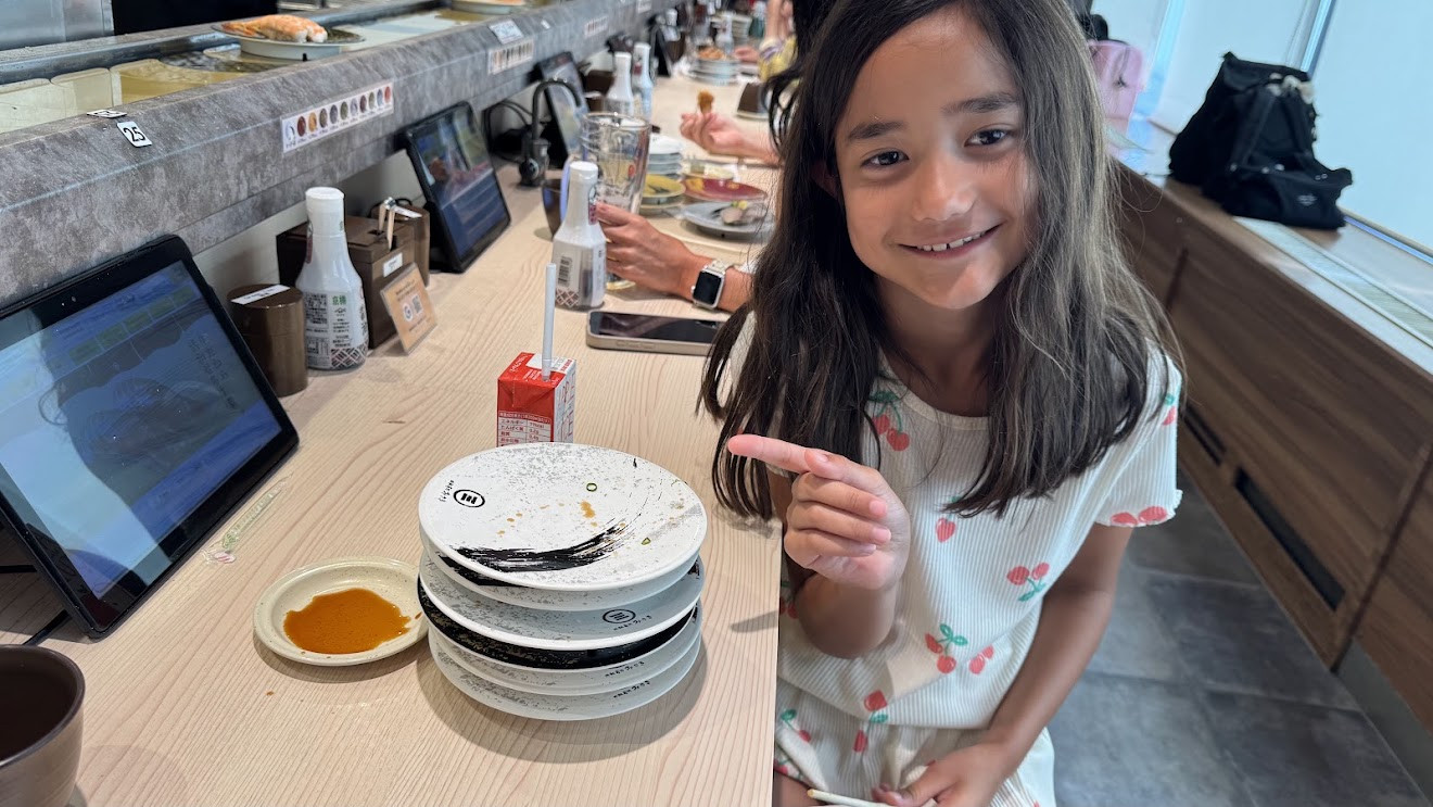 My daughter smiling and pointing at her stack of sushi plates at a conveyor-belt sushi restaurant