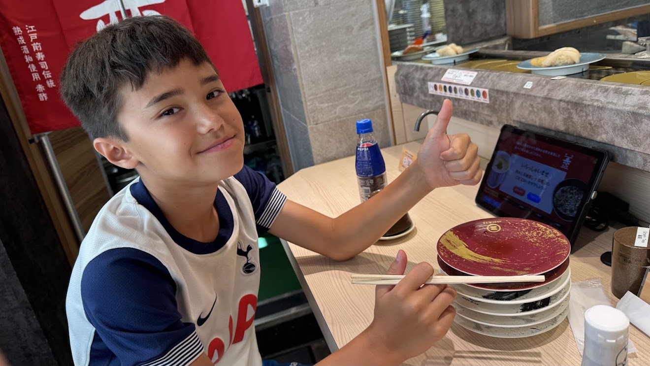 My son giving a thumbs up next to a tall stack of empty plates at a conveyor-belt sushi restaurant in Japan
