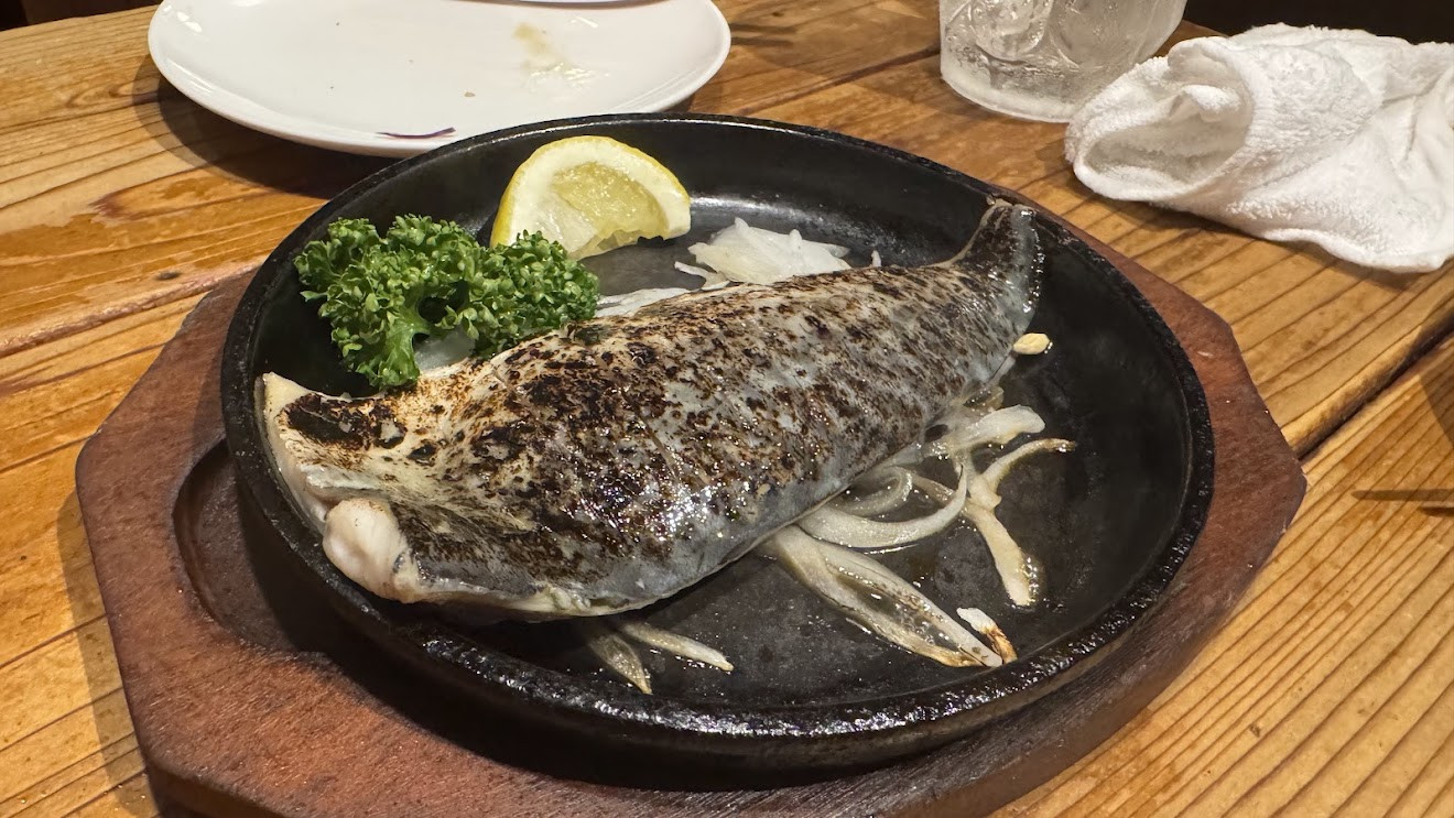 A whole grilled fish with onions, lemon and parsley on a sizzling iron plate at an izakaya
