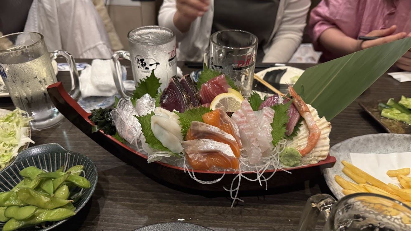 A wooden boat piled with sashimi on an izakaya table, with bowls of edamame and fries