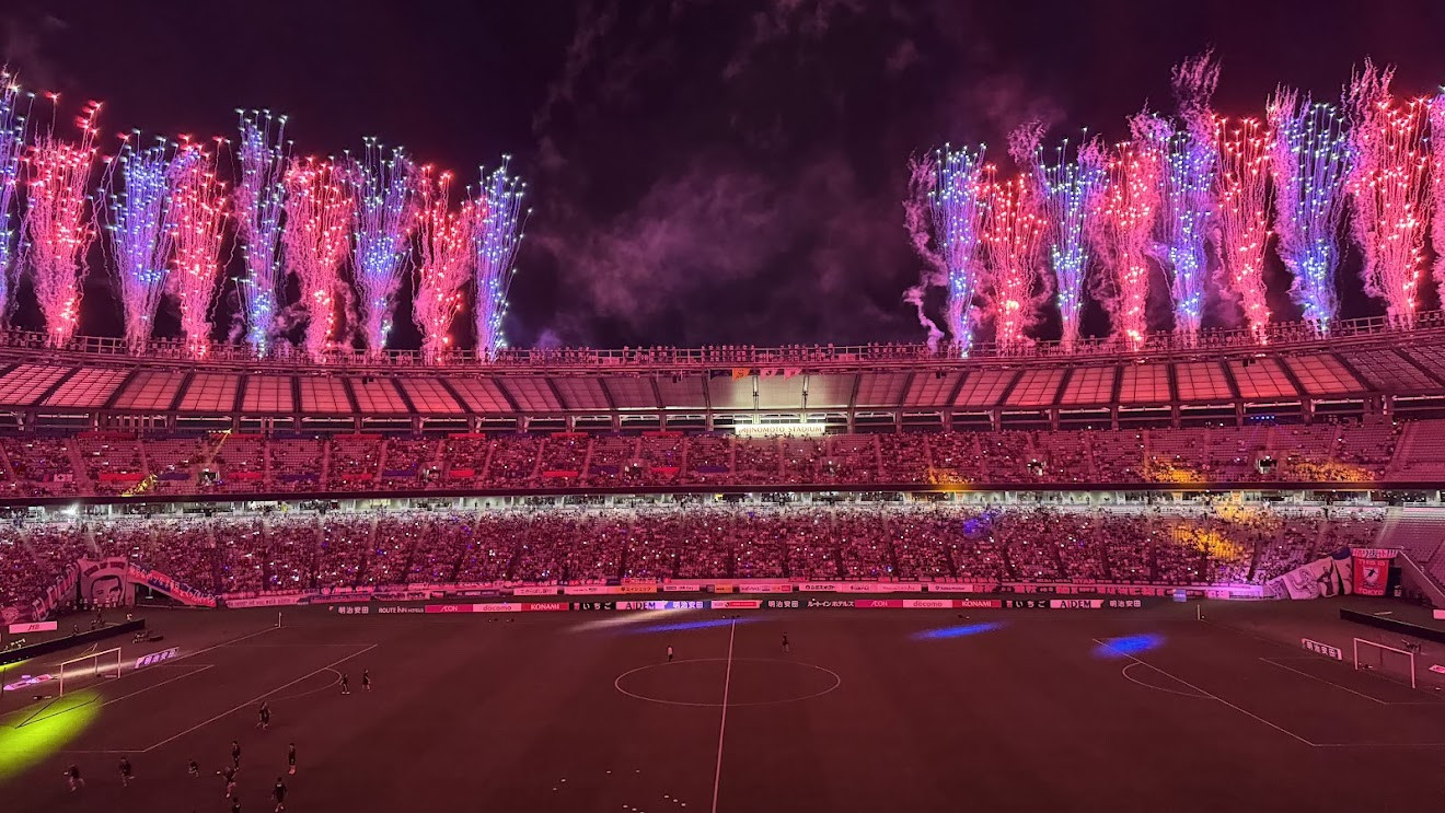 Pink and blue fireworks shooting up above a packed soccer stadium at night