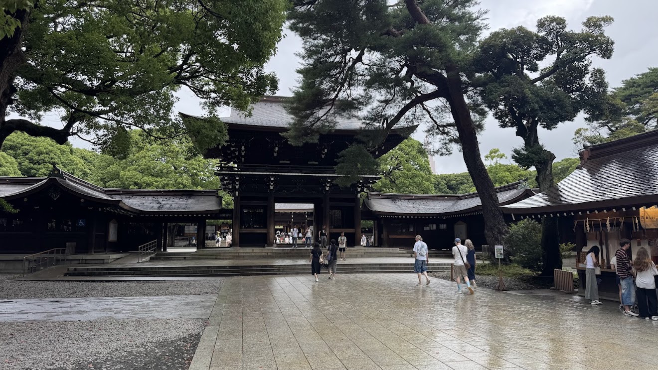 A wooden shrine gate and courtyard at Meiji Jingu on a rainy day, with wet stone paving and big trees