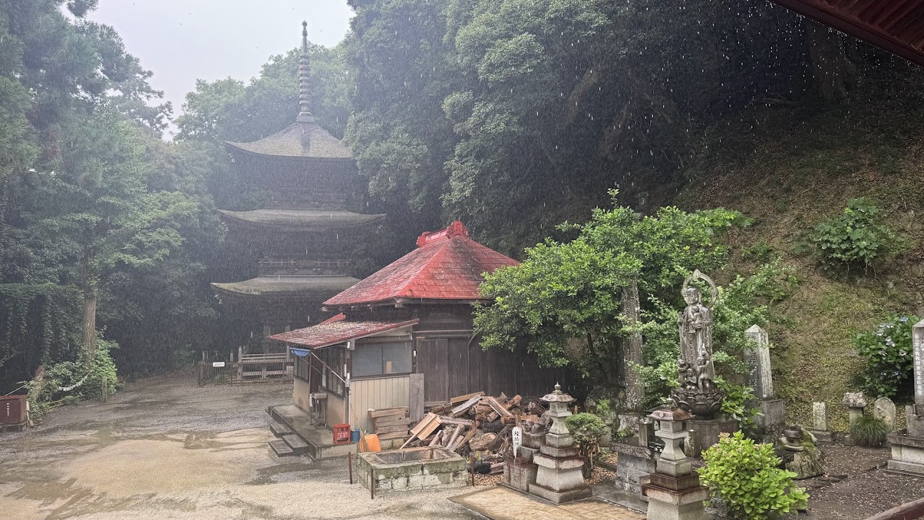 A three-story pagoda rising above a small red-roofed temple building and stone statues in a wooded hillside, in the rain